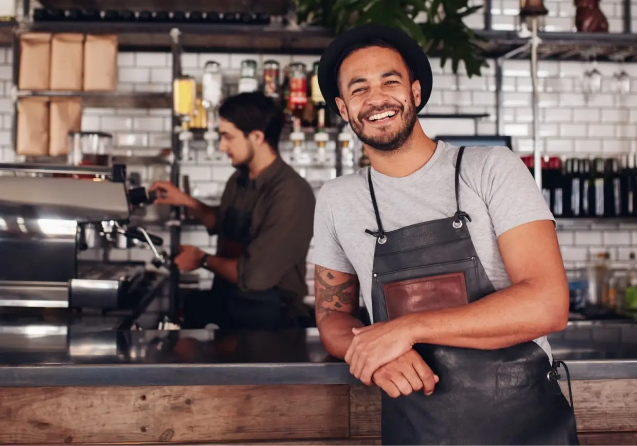 Café-Inhaber lehnt am Tresen, während ein Kollege im Hintergrund Kaffee zubereitet